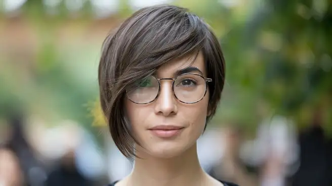 A woman with glasses showcases a stylish long pixie cut with asymmetrical bangs, highlighting her modern hairstyle.